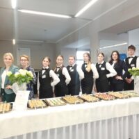 Nauczyciele i uczniowie ustawieni za stołem bankietowym z przekąskami
